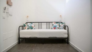 A small, narrow room with a single black metal bed frame and white bedding. There are two wall-mounted light fixtures and a small wooden shelf on the left wall. - APT. - Studio apartment in the historic center of Naples (Napoli)