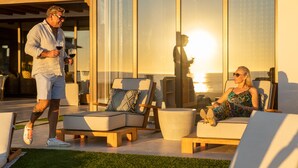 Un área de estar moderna al aire libre con sillones reclinables y un sofá, una pared de vidrio que refleja el atardecer, y una persona sosteniendo una bebida.