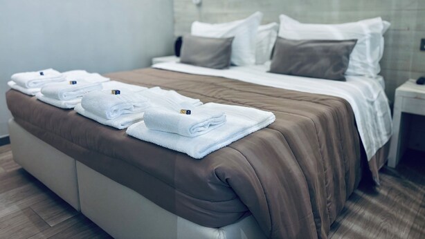 A neatly made bed with a brown bedspread and white pillows, a wooden nightstand with a lamp, and a stack of folded white towels on the bed. - Appartamento - Angolo di Rio (Riomaggiore)