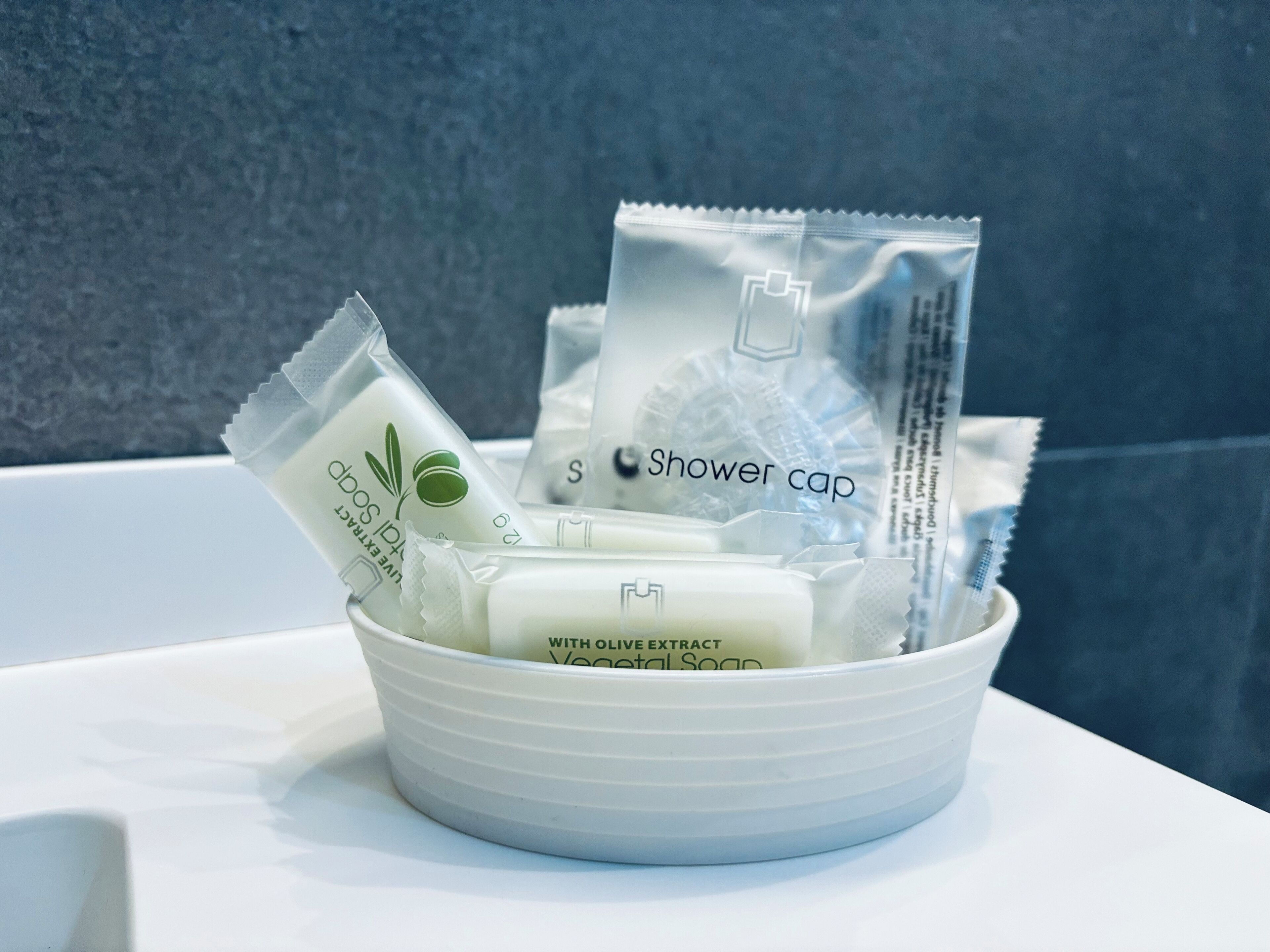 Un bol blanc contenant divers articles de toilette, notamment un bonnet de douche et un savon à l’extrait d’olive.