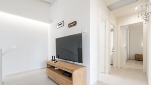 A modern living room with a large flat-screen TV on a wooden stand, white walls, and a hallway leading to other rooms. - Villetta Ada: a little corner of paradise just steps from Poetto (Cagliari)
