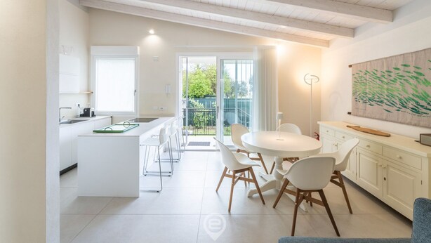 A modern kitchen with white cabinetry, a central island, and a dining area with white chairs. - Villetta Ada: a little corner of paradise just steps from Poetto (Cagliari)