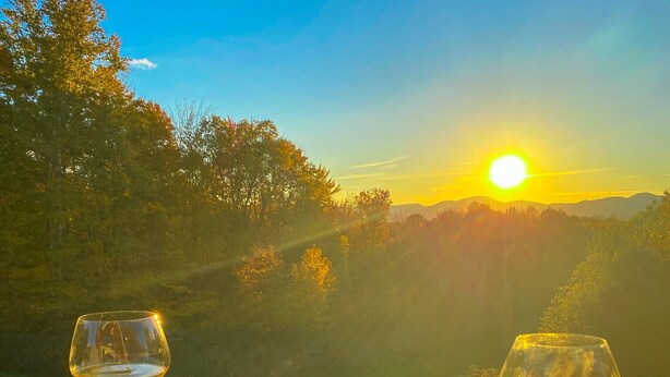 Une table avec deux verres de vin blanc et une assiette de fromage, le tout placé devant un magnifique paysage au coucher du soleil.