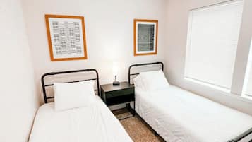 A small, clean room with two single beds, a nightstand, and a window with blinds.