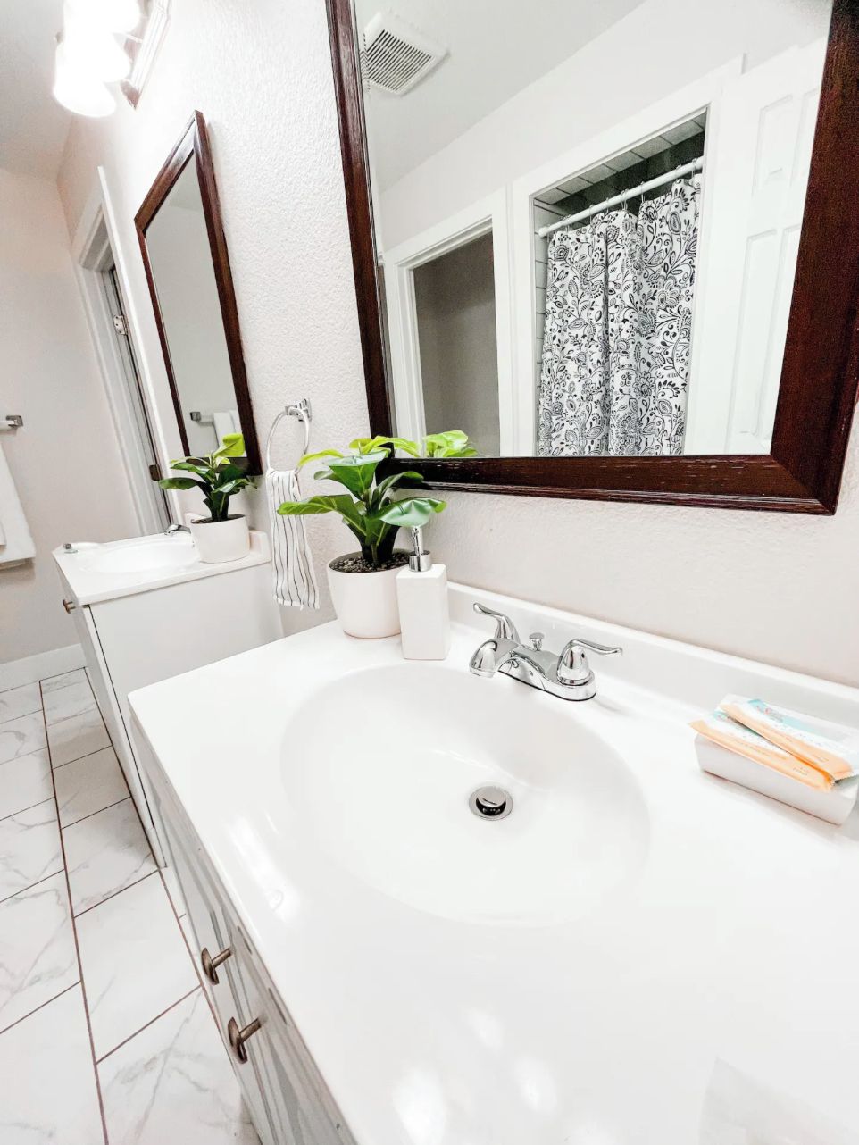 A modern bathroom with a white sink, two mirrors, a window with patterned curtains, and a potted plant.