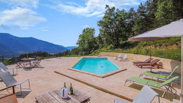 Área de piscina com espreguiçadeiras, mesa de madeira com garrafas e vista para as montanhas.