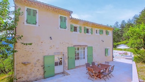 Um edifício de dois andares com telhado de telhas, venezianas verdes e um pátio com mesa e cadeiras.