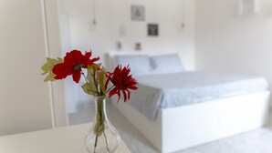 Una habitación con una cama, una mesa blanca con un florero y una pared blanca con un cuadro.