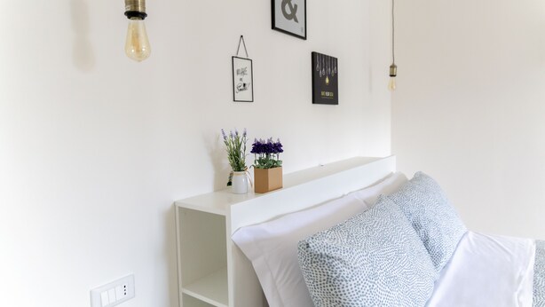 Un dormitorio con una cama blanca, una mesita de noche con un florero y lámparas fijadas a la pared.