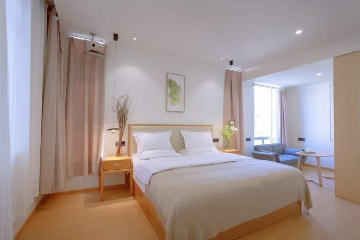 A modern hotel room with a large bed, a bedside table, a small table, and a window with curtains.