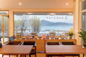 A restaurant with a view of a lake and mountains, wooden tables and chairs, and a shelf with various items.