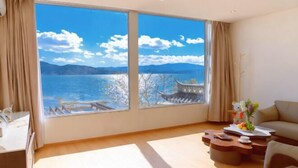 Ein Zimmer mit großem Fenster und Blick auf einen See und Berge, eine Schlafcouch, ein Couchtisch und ein kleiner Schreibtisch mit Lampe.