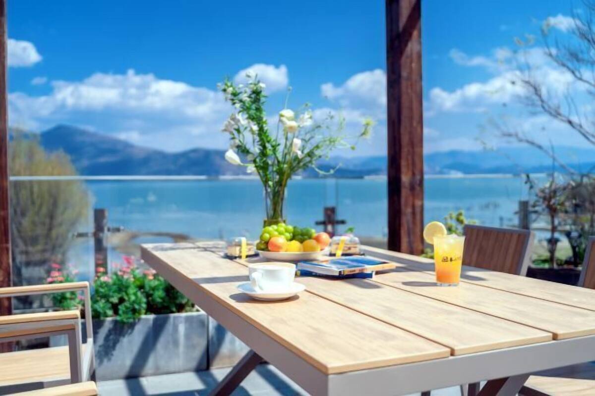 A wooden table with a view of a lake and mountains, a vase with flowers, a bowl of fruit, a cup, and a glass of orange juice.