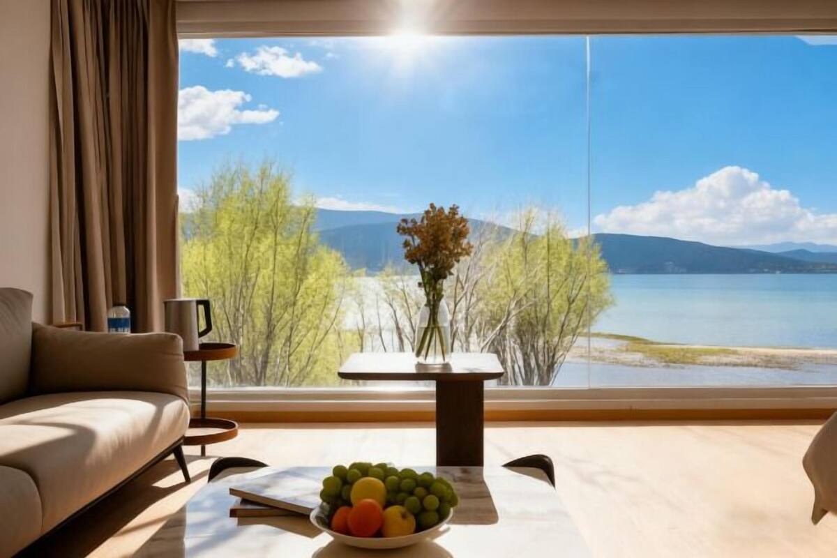 A modern room with a large window, a sofa, a small table with a bowl of fruit, and a vase with flowers.