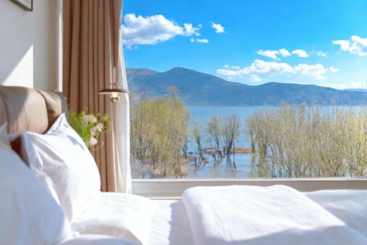 A hotel room with a bed, a window with a view of a lake and mountains, and a curtain.