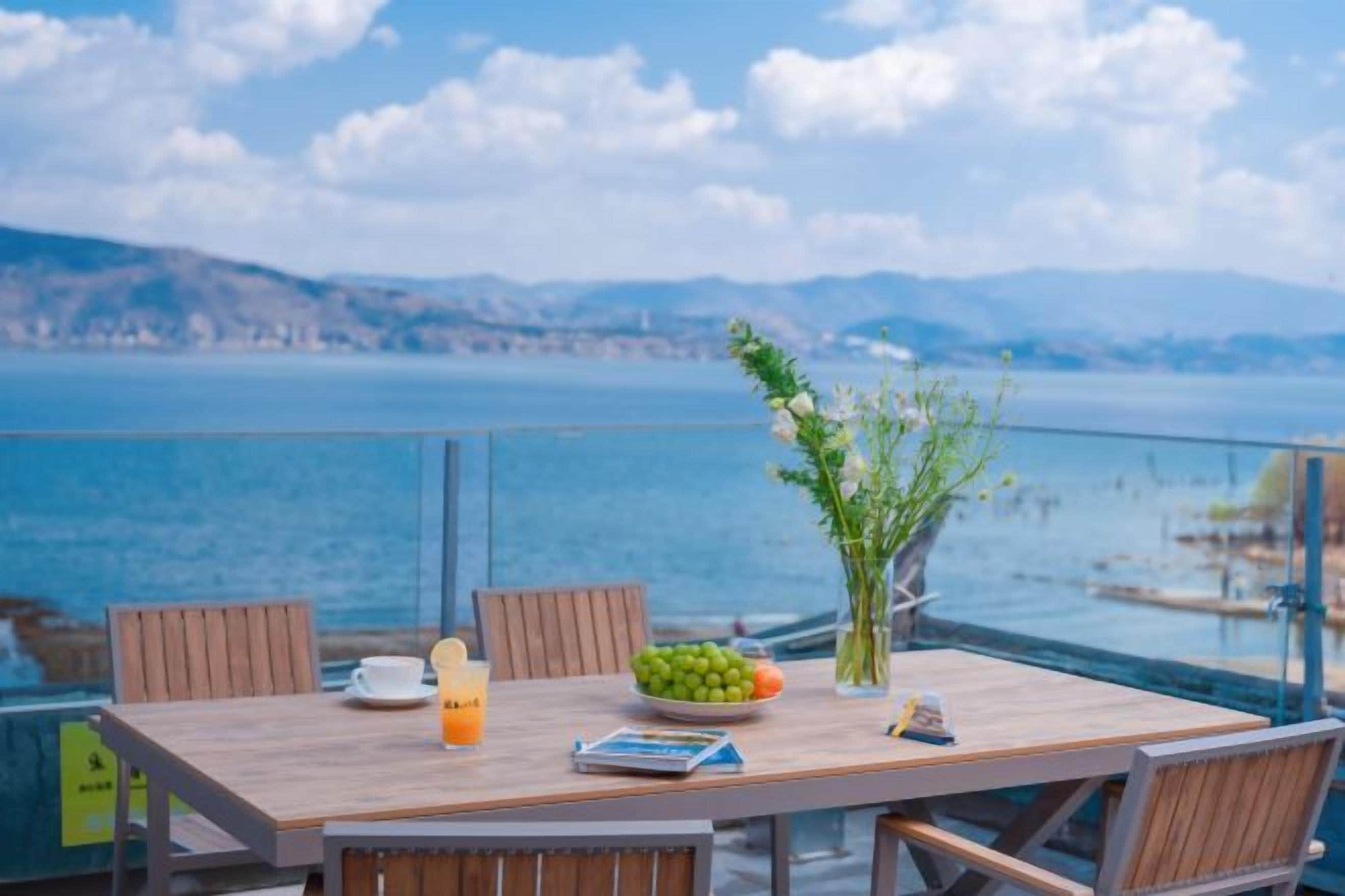 Une table en bois avec des chaises, un vase rempli de fleurs, un saladier de fruits, un verre de jus d’orange et un menu, le tout sur un balcon donnant sur une étendue d’eau et des montagnes lointaines.