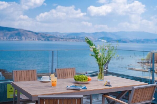 Una mesa de madera con sillas, un jarrón con flores, un cuenco de frutas, un vaso de jugo de naranja y un menú en un balcón con vistas a un cuerpo de agua y montañas distantes.
