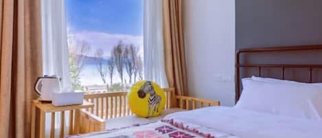 A hotel room with a bed, a wooden nightstand, a kettle, and a view of mountains through the window.