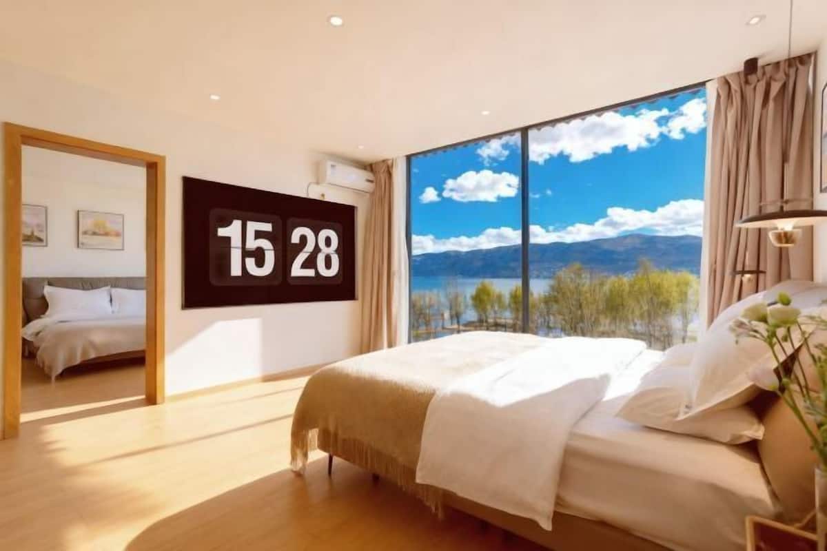 A bedroom with a large window, a bed, a wall clock, and a view of mountains and trees.
