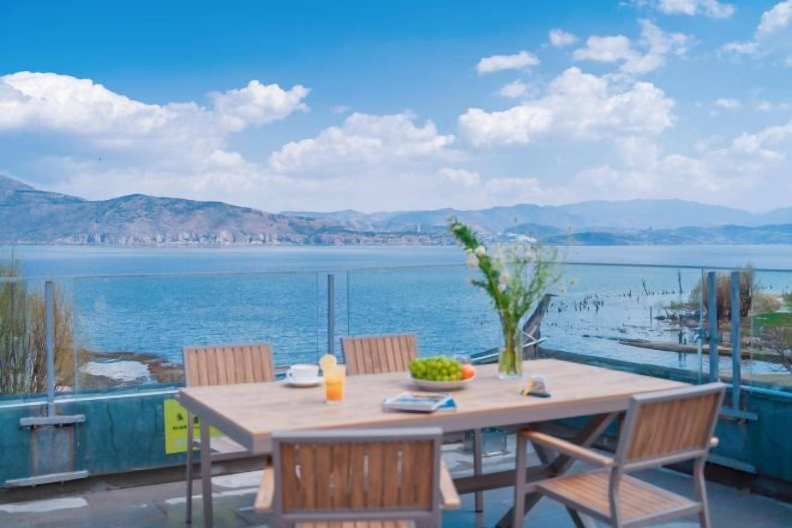 A table with chairs and a vase of flowers, overlooking a body of water and mountains.