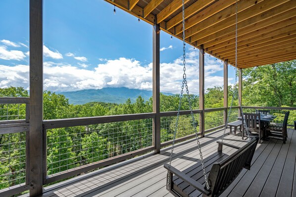 A wooden deck with a swing and a table and chairs, overlooking a forested mountain range. - HUGE VIEW | Pool | Pool Table | Theater Room (Gatlinburg)