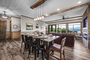A spacious dining area with a large table set for six, surrounded by wooden chairs. A cozy seating area with a brown sofa and a fireplace is visible in the background. - HUGE VIEW | Pool | Pool Table | Theater Room (Gatlinburg)
