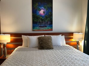 Ein Hotelzimmer mit einem großen Bett, zwei Nachttischlampen, einem Holzkopfstück und einem Wandbild mit einer tropischen Szene.