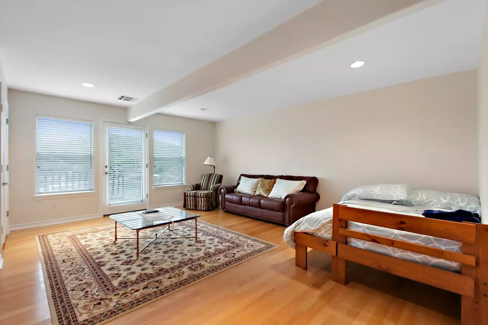Un salón amplio con un sofá de cuero marrón, una cama de madera, un sillón a rayas y una mesa de centro de vidrio.