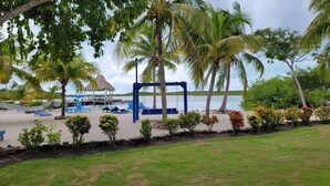 Una zona de playa con palmeras, un conjunto de columpios y una estructura con techo de paja.