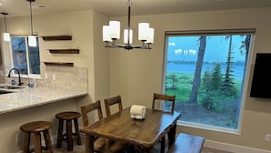 Un coin repas avec une table en bois, six chaises et un banc. Une grande fenêtre offre une vue sur les arbres et un plan d’eau.