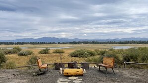 Un espace extérieur rustique aménagé avec deux bancs en bois et une assise en tronc d’arbre, le tout entouré d’un paysage naturel avec des montagnes en arrière-plan.