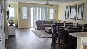 A spacious living room with a dark hardwood floor, a beige sofa, a dining area with a round table, and a kitchen area with a countertop.