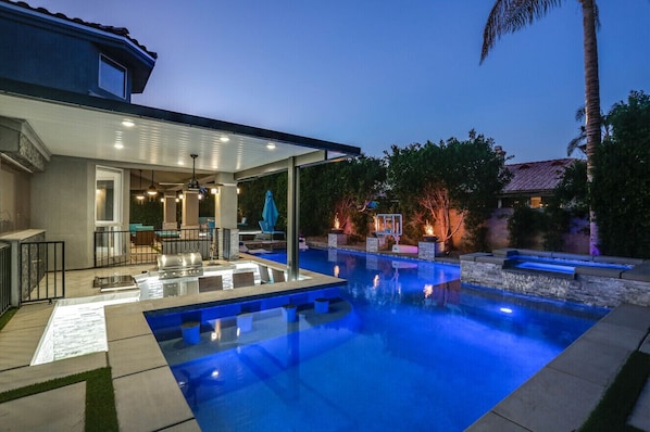 A modern outdoor pool area with a covered patio, barbecue grill, and a basketball hoop.