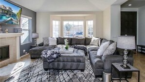 Un salon moderne avec un canapé d’angle, une cheminée et de grandes fenêtres donnant sur l’extérieur.