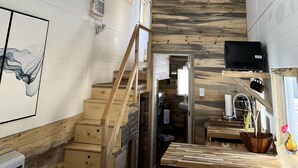 A rustic interior with exposed wooden beams, a ceiling fan, a staircase, a kitchen area, and a mounted television. - The Luxe Tiny Cottage  (Mayhill)