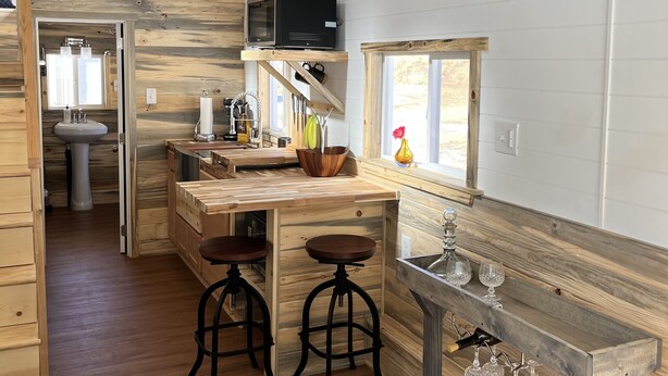A rustic kitchen with wooden walls, a wooden countertop, and a built-in microwave. There are two stools, a sink, and a window with a view. - The Luxe Tiny Cottage  (Mayhill)