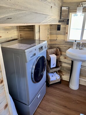 A laundry area with a front-loading washing machine, wicker baskets, and a sink with hand soap. - The Luxe Tiny Cottage  (Mayhill)