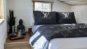 A bedroom with a bed, two pillows with the Louis Vuitton logo, a Buddha statue, a glass decanter, and a tray with glasses. - The Luxe Tiny Cottage  (Mayhill)