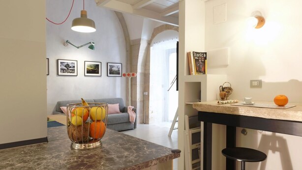 Een keuken met een marmeren aanrecht, een fruitschaal, een hanglamp en een boekenkast met een boek getiteld 'Storia'.