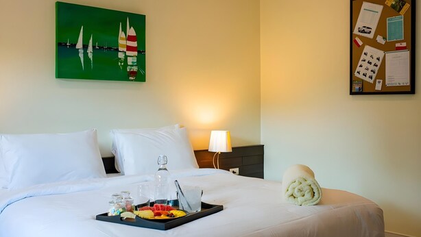 A neatly made bed with white linens, a tray of fruit and a bottle, a painting of sailboats, and a bulletin board with papers and photos. - The Lokal Hotel Phuket (Patong)