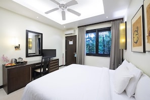 A hotel room with a bed, a desk with a chair, a television, a ceiling fan, a window with curtains, and a framed picture on the wall.