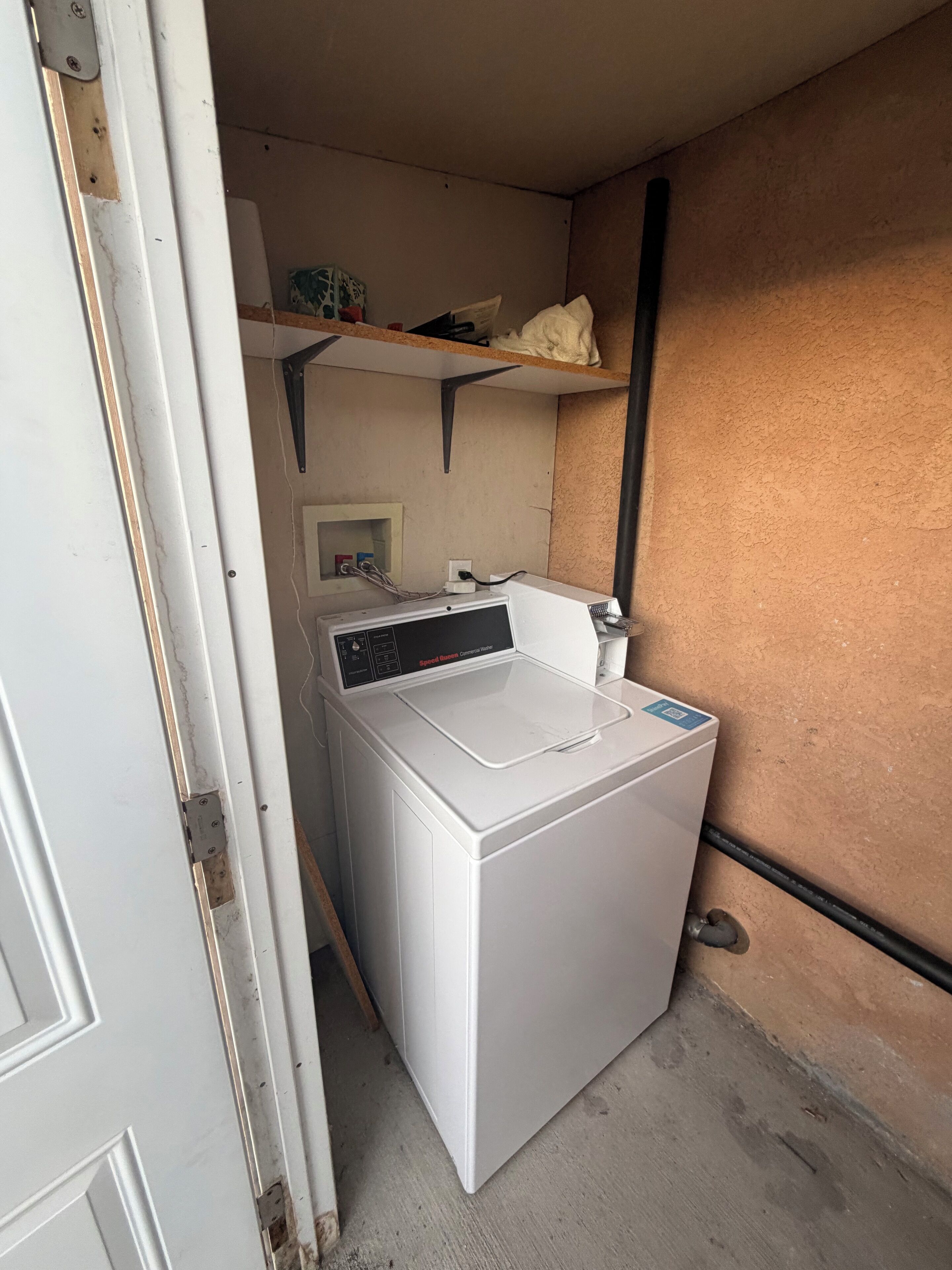 A compact laundry room with a washing machine, a shelf above, and a small electrical outlet.