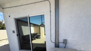 Fachada de un edificio con puerta de vidrio corredera, unidad de aire acondicionado Mitsubishi eléctrica y un pequeño espacio exterior con piso de concreto y un tapete oscuro.