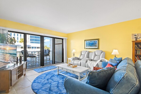 A living room with a sofa, armchairs, a coffee table, and a TV. There is a sliding glass door leading to an outdoor area with a pool and chairs.
