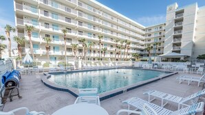 Zona de piscina de hotel con una pileta grande, sillas de descanso y un edificio con varios balcones.