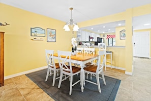 A dining area with a wooden table and white chairs, a kitchen in the background, and artwork on the walls.