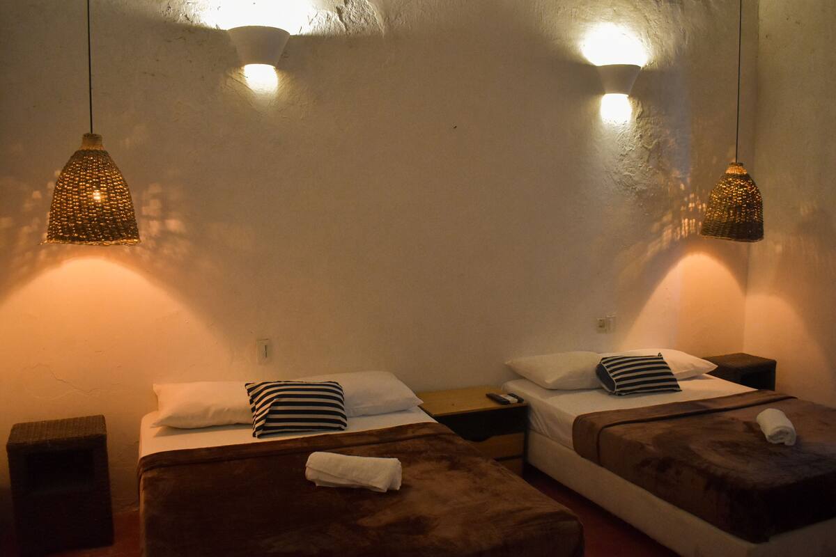 Une chambre d’hôtel avec deux lits, chacun agrémenté d’un oreiller à rayures et d’une tête de lit marron. Il y a deux tables de chevet, dont l’une porte une télécommande. La chambre présente un design simple et épuré, avec un mur blanc et un plafond texturé.