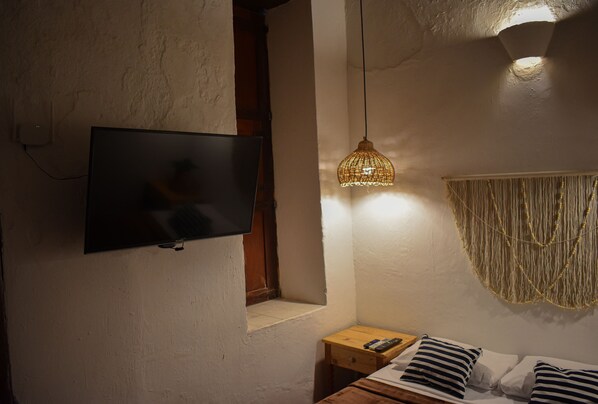 Un dormitorio con una televisión montada en la pared, una mesita de noche con un control remoto, una cama con almohadas a rayas y una lámpara de fibra trenzada.
