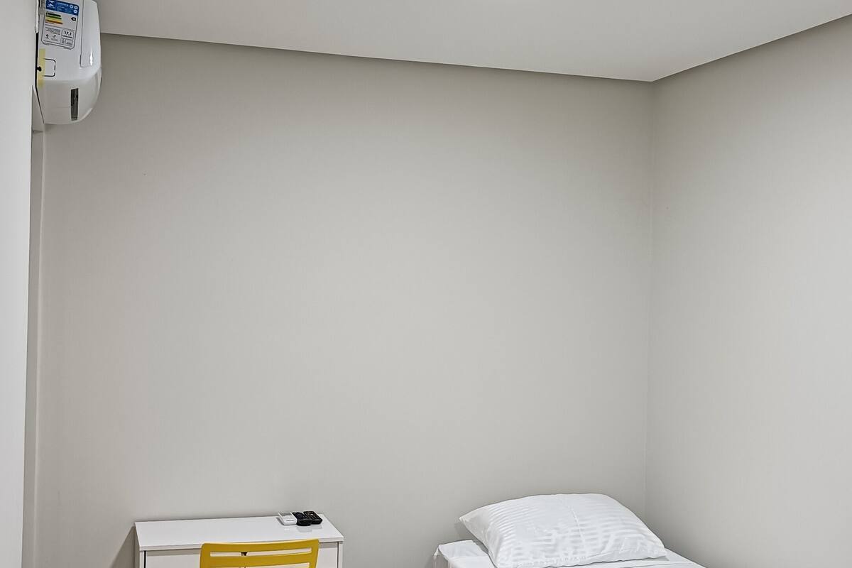 Ein kleines, sauberes Zimmer mit einem weißen Schreibtisch, einem gelben Stuhl, einem Einzelbett mit grüner Decke und einem an der Decke angebrachten Rauchmelder.
