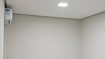 Una habitación pequeña y limpia con un escritorio blanco, una silla amarilla, una cama individual con una manta verde y un detector de humo en el techo.